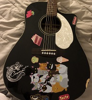 black acoustic guitar with a white pickguard, with some fun stickers.